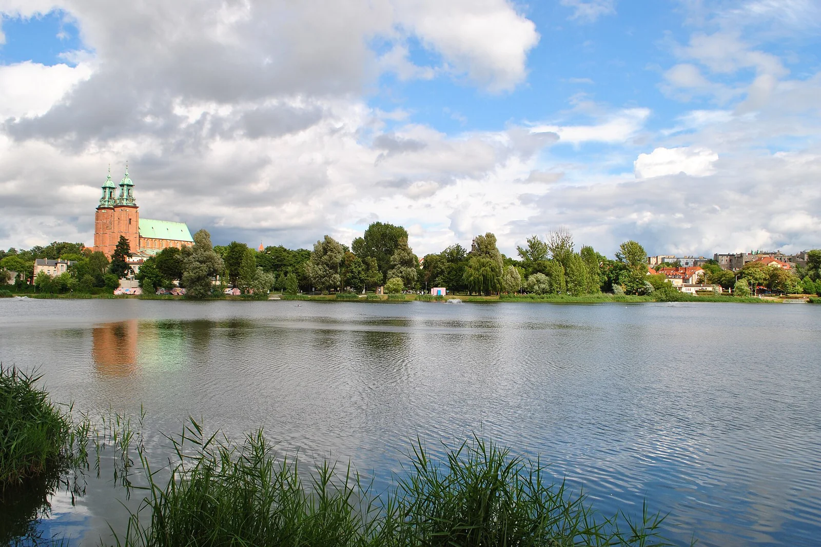 Szeroki widok Jeziora Jelonek w Gnieźnie, katedra po lewej i zieleń nad brzegiem pod rozświetlonymi chmurami