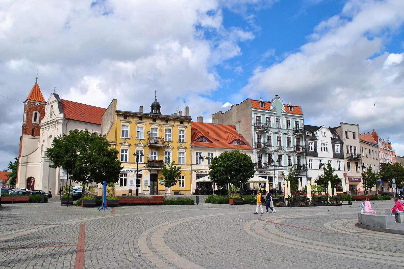 Kolorowe fasady północnej pierzei Rynku w Gnieźnie, drzewa i brukowany plac pod niebieskim niebem