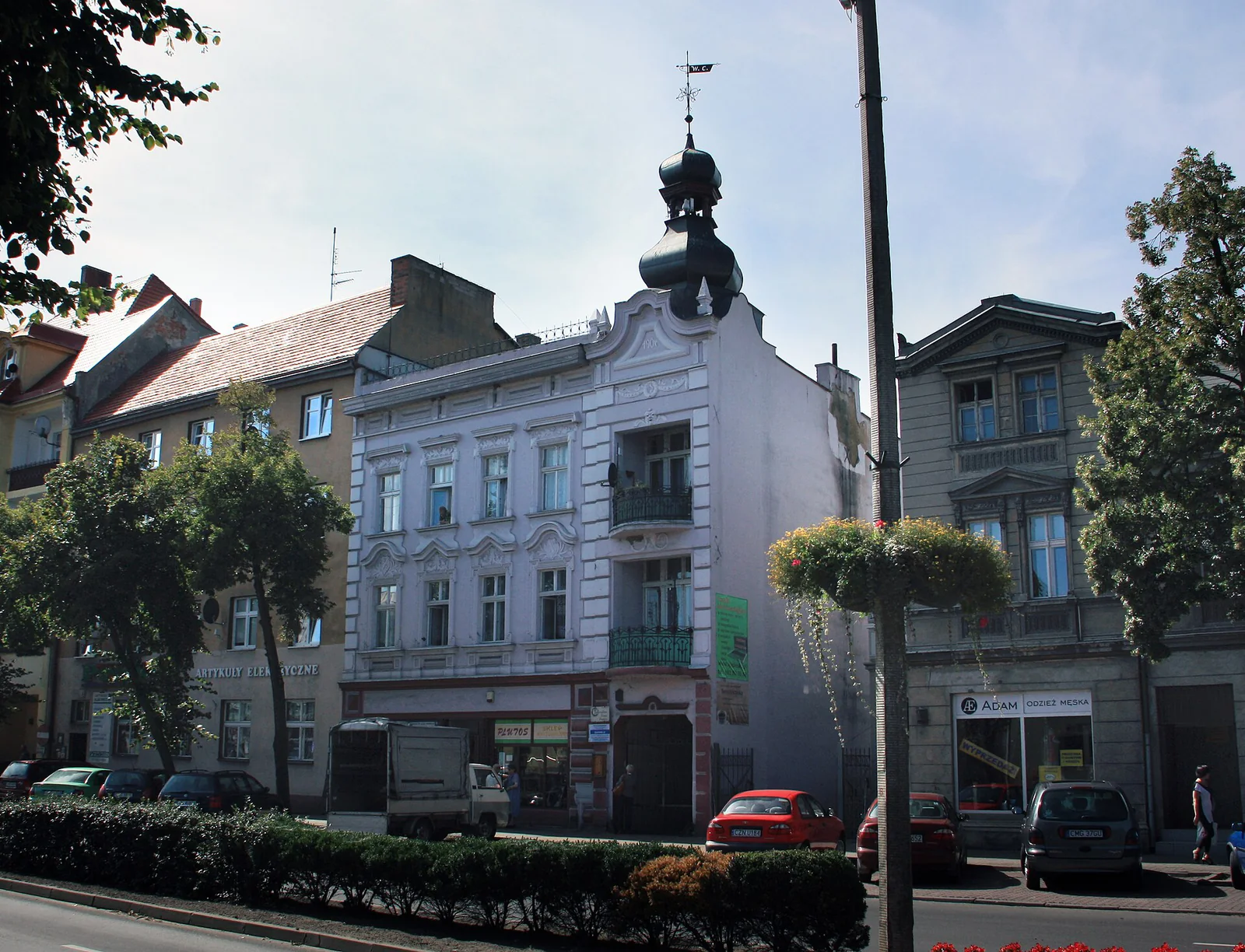 Jasna ozdobna fasada kamienicy przy placu Wolności 8 w Mogilnie, z wieżyczką i drzewami przed budynkiem