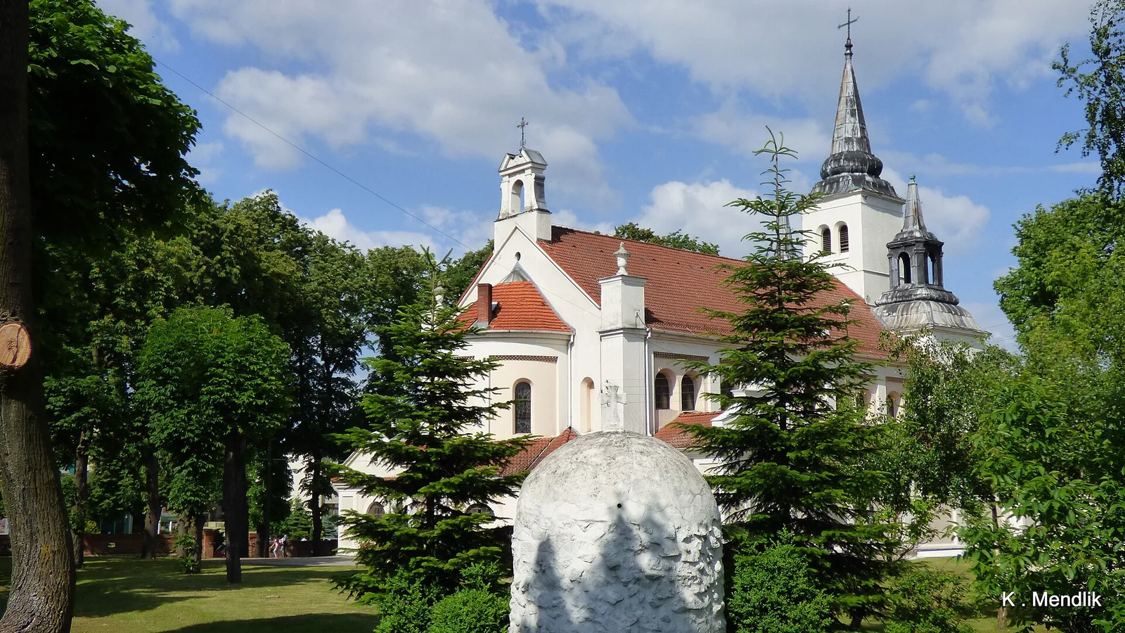 Kościół świętego Wawrzyńca w Nakle nad Notecią za fontanną i zielonymi drzewami pod błękitnym niebem