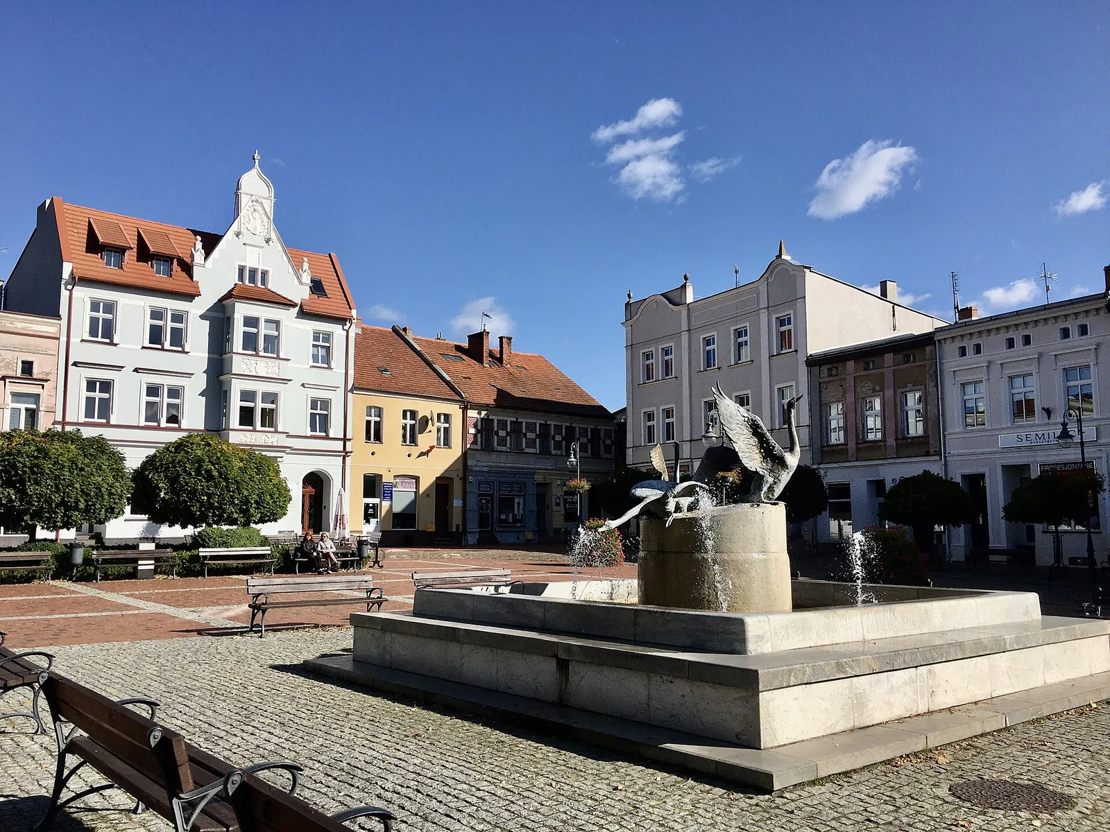 Rynek w Tucholi z fontanną, ławkami i kolorowymi kamienicami na tle błękitnego nieba